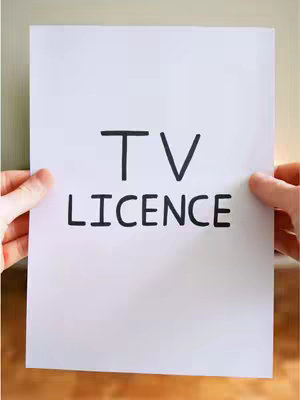 A person holding a white paper with the words "TV LICENCE" written in large black capital letters. The background is a light, neutral color, and the hands holding the paper are visible, implying the person is facing the camera.