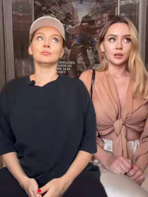 Two young women are seated in an indoor setting. The woman on the left is wearing a white baseball cap and a black oversized t-shirt, with her gaze directed upwards. The woman on the right is wearing a light brown top with spaghetti straps and is looking towards the camera. Behind them is a partially visible poster.