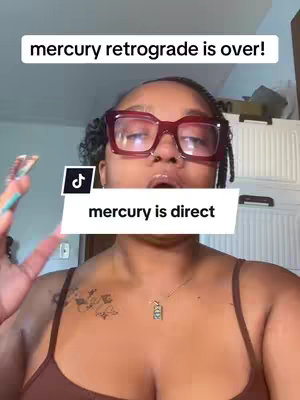 A selfie-style video cover featuring a person with dark hair, wearing large, square, red-rimmed glasses. They are looking up toward the top of the frame with slightly opened lips. Overlayed text at the top reads "mercury retrograde is over!", and another text overlay at the bottom saying "mercury is direct". She is wearing a brown tank top and has a necklace with a small object.