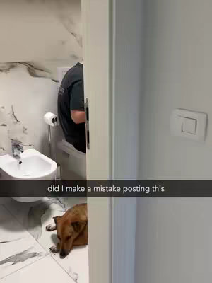 A bathroom scene is partially revealed through a doorway. A person in a dark t-shirt is sitting on the toilet. A dog lies on the tiled floor in the foreground, with its head resting on its paws, appearing relaxed. The walls have a marble design and the whole scene is captured in a candid style.