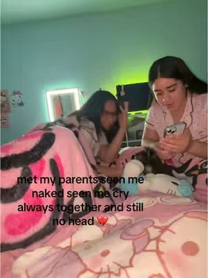 Two young women are in a bedroom setting, one appears to be using her phone while the other lies in bed under a pink blanket and comforter. Text overlay appears on this colorful, slightly blurred image.
