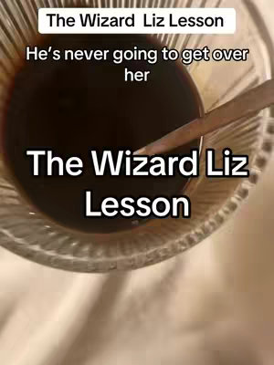 The image shows a close-up view from above a clear glass filled with dark liquid, likely coffee or tea. A spoon rests in the glass. Text overlays are present, composed of a white font with a black outline and a black background, indicating emphasis on “The Wizard Liz Lesson.” The composition focuses on the beverage and the descriptive text surrounding it.