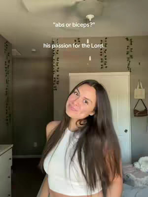A woman in a white halter top smiles towards the camera. The background suggests an interior space, possibly a bedroom, with vines hanging on the wall. The lighting is natural, highlighting her face. Text overlays are present.