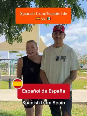 Two people stand outside, the woman on the left smiling with her arm next to the chest of a man on the right, both looking directly at the camera. The background appears to be a park with a tent-like structure and a colorful playground is visible. Two text overlays are on the image, one at the top and another at the bottom with flags. The foreground features lush green grass.