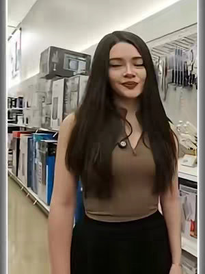 A woman with long dark hair is standing in a store aisle filled with shelves of products. She is wearing a brown top and black skirt. She is looking towards the camera with a slight smile.