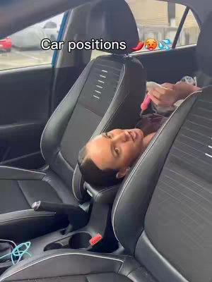 A woman is lying down in the front seat of a black car, her head resting on the armrest between the seats. The car's interior is visible, with black leather seats and a blue exterior visible through the window.