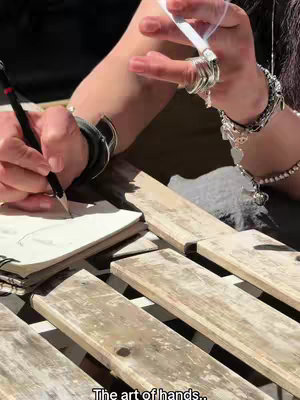 The image shows a person's hands, holding a lit cigarette and a pencil, positioned over a wooden surface. The person is wearing multiple rings and bracelets, and their other hand holds a pencil over a notebook. The setting appears to be outdoors, with sunlight casting shadows.