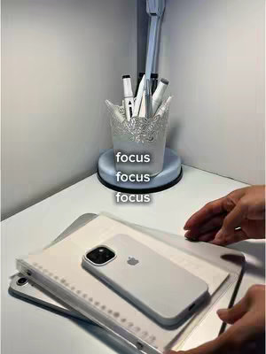 A well-lit shot of a desk with study supplies. A glass holder with pens and pencils sits on a round base, a white phone lays on top of a notebook stack and an iPad, hands appearing on the right side.