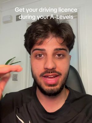 A selfie-style shot featuring a young man with dark hair and a beard centered in the frame. Overlaid text in a semi-transparent box reads "Get your driving licence during your A-Levels", positioned at the top. The man looks directly at the camera, conveying a conversational tone. The background shows a simplified neutral wall, a door, and a plant, suggesting a home setting.