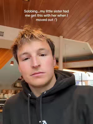 A selfie-style shot featuring a young man with short, messy light-brown hair and a neutral expression. He's wearing a black hoodie, which is partially obscuring his shoulders. The background is somewhat blurred, but shows hints of a bright, airy interior with sections of wooden panels and what appears to be a countertop with coffee-related items. Text overlayed on the image reads "Sobbing...my little sister had me get this with her when I moved out :')", The lighting appears soft with warm tones.