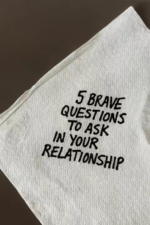 The image is centered on a white, creased paper napkin. Bold, black handwritten text dominates the napkin, reading "5 BRAVE QUESTIONS TO ASK IN YOUR RELATIONSHIP". The text is arranged in a stacked format, with each line centered. The napkin is angled, and the background is a solid, dark brown.