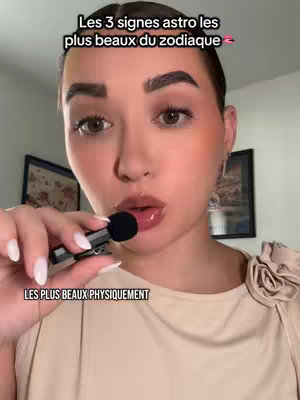 A young woman with light skin is centered in a selfie-style shot. She has a serious expression with her lips slightly parted, and she is holding a small black microphone close to her mouth. The text overlay at the bottom reads "LES PLUS BEAUX PHYSIQUEMENT". The background is softly blurred, including two framed pictures with indiscernible images.