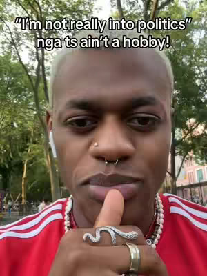 A medium shot shows a person with light blond, short hair looking straight ahead. The image is framed from the chest up. The person is wearing a red shirt with white stripes on the shoulder, paired with a pearl necklace. Facial features include a nose ring and a septum piercing. They are holding up their pointer finger to their lips. On the finger is a silver ring shaped like a snake, a golden ring and a silver ring. The background appears to be a city park.