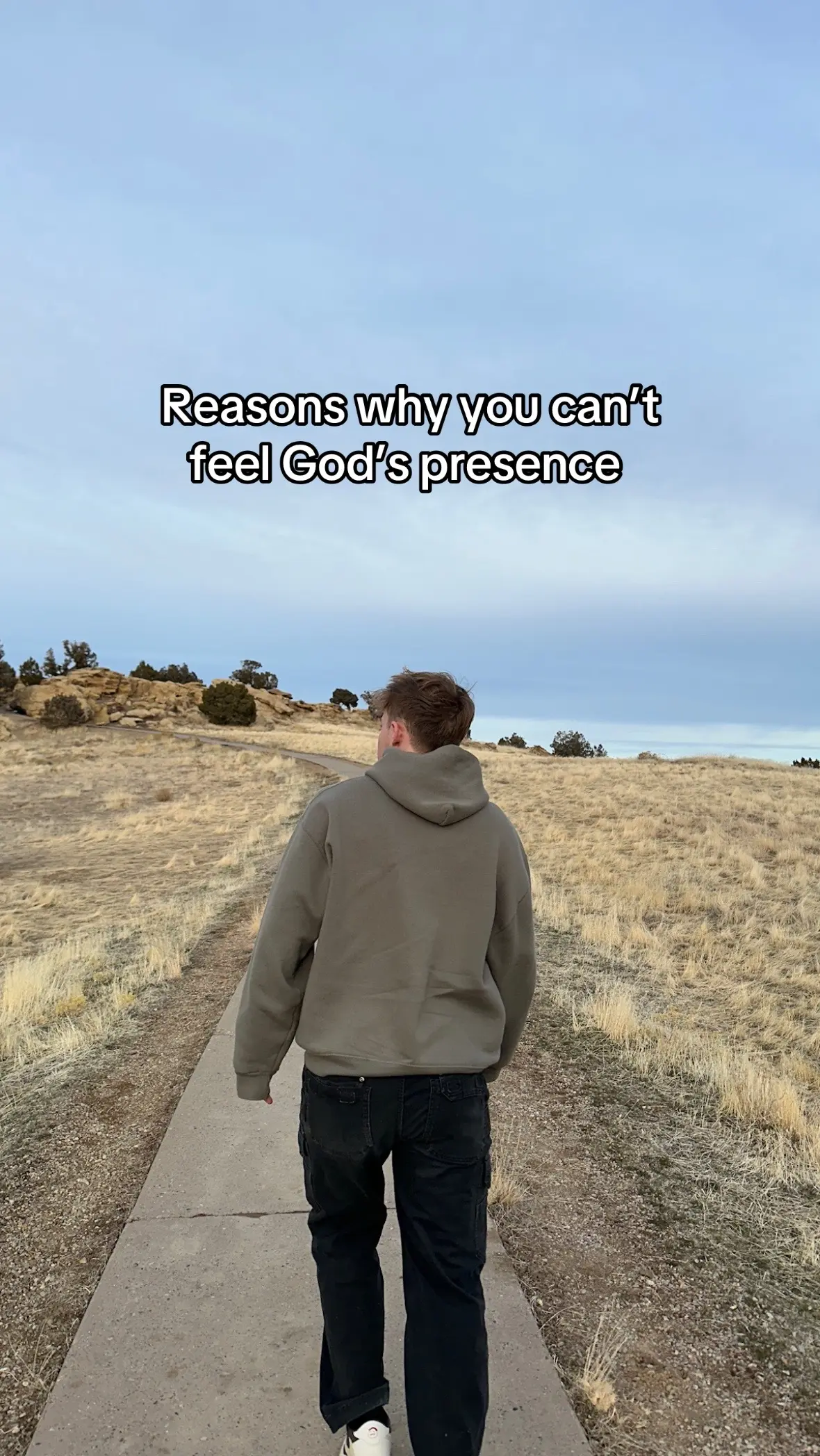 A person is walking away from the camera on a concrete path. They are wearing a grey hoodie, black pants and white sneakers. The path leads through an area of dry, yellow grass and low bushes under a slightly overcast sky. Text is overlaid at the top, in white letters.