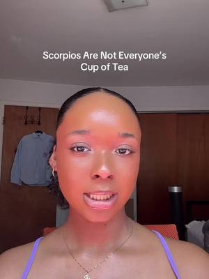 A selfie-style shot featuring a woman with light makeup looking directly at the camera, framed from the chest up. The background is a room with indirect lighting. The top text overlay says, "Scorpios Are Not Everyone's Cup of Tea". She is wearing a purple top and a gold necklace with a pendant.