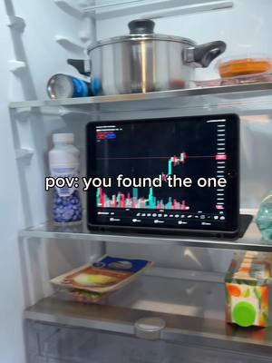 A well-lit shot from inside a refrigerator shows various items on shelves. The main focus is on a tablet displaying a stock chart, labeled with the text "pov: you found the one." Other items include a cooking pot, drink cans, bottles, food packages, and juice.