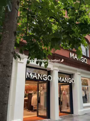 An exterior shot of a Mango store. The storefront has large glass windows, reflecting the outside. The Mango logo is prominently displayed above the entrance. The image is partially obscured by tree branches, suggesting an outdoor setting.