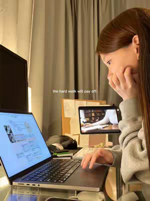 A young woman is seated at a desk in a room, illuminated by warm lighting. She is facing towards the right, with her head leaned down, her right hand covering her mouth, and her eyes looking towards the screen. She is surrounded by office supplies and two screens; one is a laptop displaying a website or document, the other is a tablet showing some writing. The photo gives off a studious, focused feeling. A text overlay at the bottom reads "the hard work will pay off."