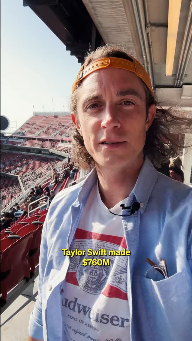 A man in a yellow cap and a light blue button-down shirt is standing in front of a stadium full of people. The stadium has red seats.