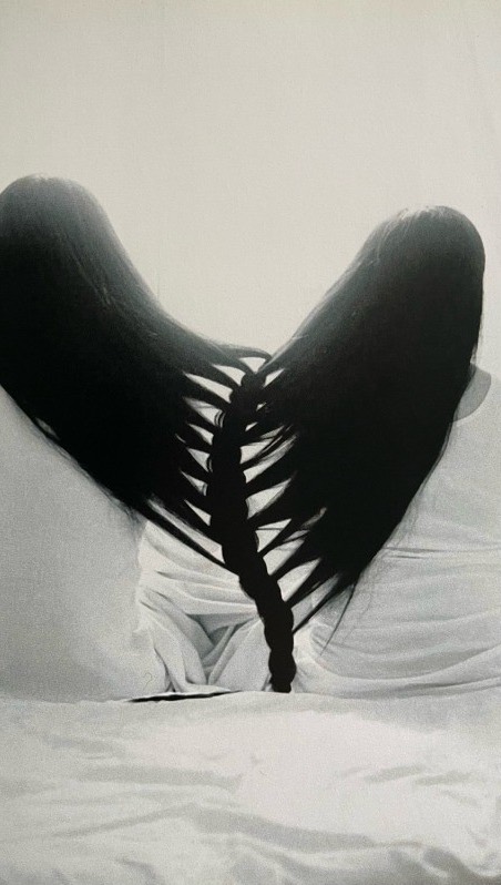 A black and white image featuring two figures, likely women, seen from the back. Their long, dark hair cascades down, forming a heart shape with a braid at the center where the hair meets. They appear to be sitting or reclining, with their upper bodies and hair taking up most of the frame. The background is plain and neutral. The contrast between the dark hair and light background is stark.