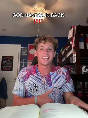 A young man with short, light-brown hair sits in a room with a ceiling fan and overhead light. The hook text "GOD HAS YOUR BACK" is at the top center. He is smiling, wearing a patterned shirt, and pointing his finger. An open book is on the table in front of him. The background appears to a bedroom with decorations and a bunk bed.