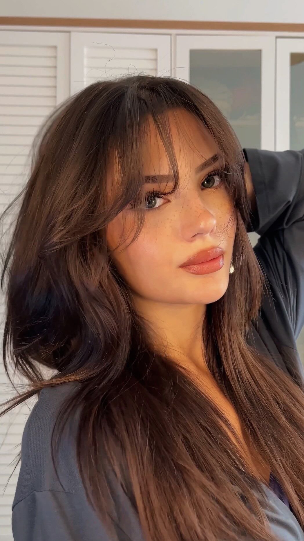 A close-up shot of a woman with long brown hair featuring subtle waves and side-swept bangs. She has freckles, full lips with light pink lipstick, and is wearing a gray top. The background is a blurred interior with what appears to be a white cabinet.