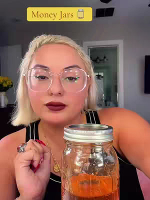 A woman with blonde hair and glasses looks directly at the camera while holding a mason jar filled with liquid. The jar is in the foreground. A text overlay at the top says "Money Jars".
