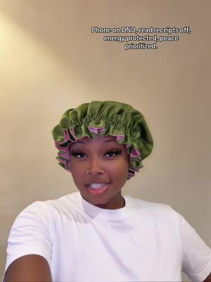 A woman with dark skin and brown eyes is smiling at the camera, wearing a green satin bonnet with a pink trim and a white t-shirt.