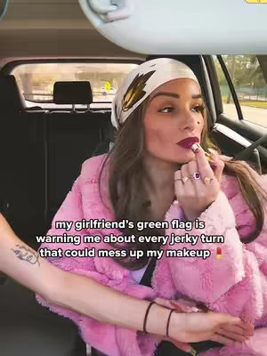 A woman in a pink fuzzy jacket applies lipstick inside a car. The camera angle is from inside the car with her arm and the arm of another person visible.. The background shows the interior of the car and the outside environment through the windshield and side window.