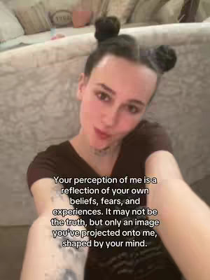 A selfie-style shot features a young woman with dark hair styled into two buns. She's looking directly at the camera, with a slight smile and a soft focus. She's wearing a dark brown top and has a delicate necklace and small tattoo. The background includes a couch in a cozy setting. Overlaying the main image is text with a thought-provoking quote touching on the perception and truth of others. The overall color palette is natural and warm due to the skin tone and the muted background.