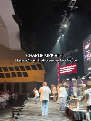 The image captures a live worship event. The stage is well-lit, featuring musicians and a singer near the front. In the foreground, a person in a white shirt is seen from the back, likely part of the worship team. The background showcases a large screen with visible text and the venue's architecture, suggesting a church setting.