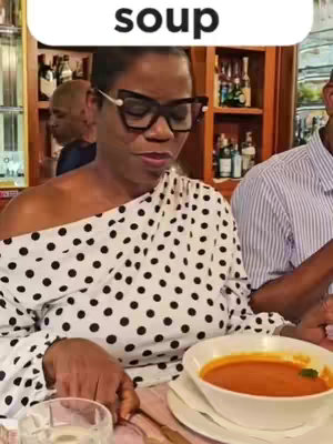 A woman wearing glasses and a polka-dot shirt is seated at a table, preparing to eat soup from a white bowl. The background shows a warm, restaurant setting with bottles of wine visible on shelves.