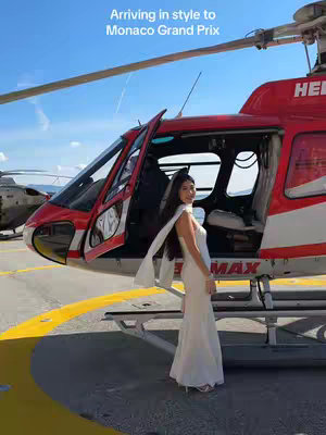 A woman in a white gown poses next to an open-doored red helicopter on a tarmac. The backdrop is a bright blue sky.