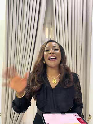 A woman with long, dark brown hair stands indoors in front of a window, looking upwards with an open mouth as if speaking excitedly. She wears a black, patterned long-sleeved top, a gold necklace with a round pendant, and has gold earrings and a bracelet. She is holding a tablet, and a curtain is the backdrop.