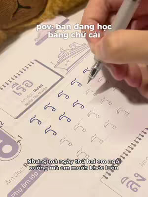 A person is seen writing characters in a notepad, using a pen. The notepad displays the Thai alphabet. There is text overlayed on top, with the words 'pov: bạn đang học bằng chữ cái' at the top. Below, a hand is writing Thai characters in the form of squiggles. At the bottom of the image, there is more overlaid text. The image has a notebook on the table and hands holding the pen, and the lighting is bright.
