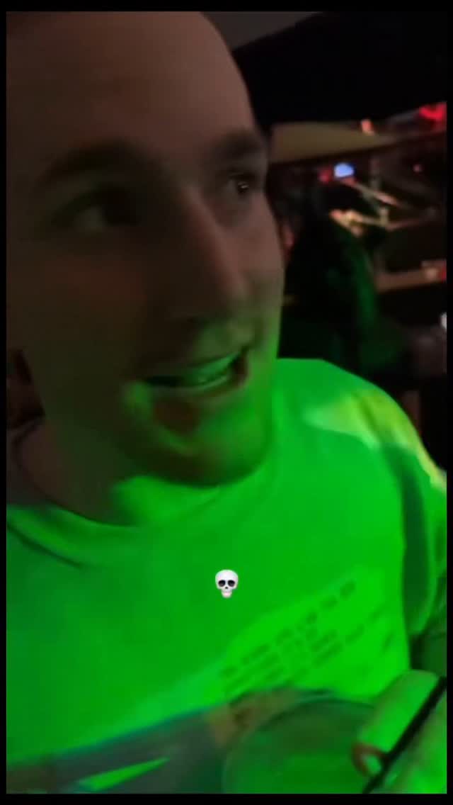 The image shows a man in close-up, slightly angled to the right, against a green background. He's wearing a green shirt, and the overall lighting gives the scene a green tint. A small skull emoji appears in the background to the right corner. The man appears to be speaking, mouth open. The composition focuses intently on his facial expression.