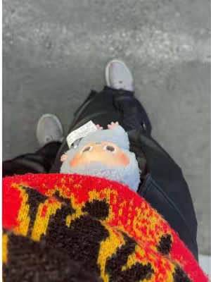 The image is taken from a high-angle perspective, showing the lower body and feet of a person, with the feet clad in gray athletic shoes. The person is seated with a plush toy resting on their lap. The upper part of a patterned red, yellow, and black sweater is also visible. The background is a smooth gray concrete surface. The composition is simple, focusing on the textures and colors of the clothing and toy against the neutral background.