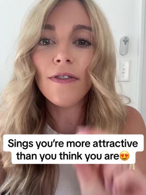 A close-up shot of a woman with long, wavy blonde hair, looking directly at the camera. She has fair skin and is wearing a white top. Text overlayed on the image reads, 'Signs you're more attractive than you think you are.' The background is a light-colored wall with a switch visible.