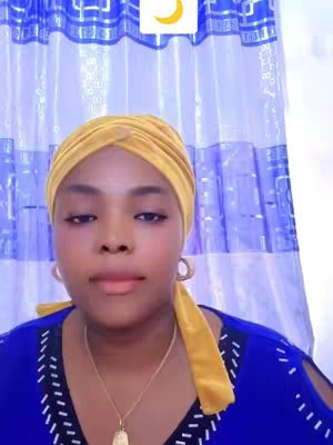 A woman with dark skin wearing a yellow head wrap and a blue top is looking directly at the camera. Behind her is a white curtain with a blue pattern. In the top center of the frame is a box containing a crescent moon graphic.