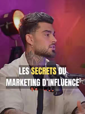 A man with tattoos is in the center of the shot, addressing the camera. He's wearing a white shirt. The backdrop features a dramatic lighting setup with a large spotlight. The image is dominated by warm tones of orange and purple.