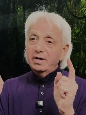 A medium shot of an older man with white hair is gesturing with his right index finger raised. He has a neutral expression and is wearing a purple shirt. The background is blurred, featuring green foliage.