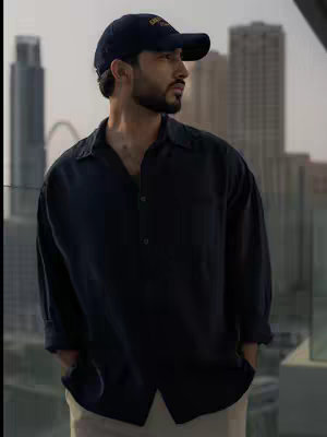 A man stands outdoors looking out at a cityscape. He wears a baseball cap, dark button-up shirt, and light-colored pants. The background shows a tall, modern city skyline under an overcast sky.