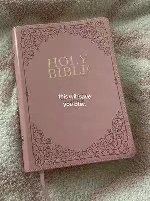 A close-up shot of a light pink Bible lying on a textured, multi-colored surface, likely a blanket or bedspread. The Bible is closed with a pink cover and has a decorative border around the edges and the title "HOLY BIBLE" embossed in a lighter color. A white ribbon is attached to the spine, serving as a bookmark. Below the title, a text overlay says "this will save you btw."