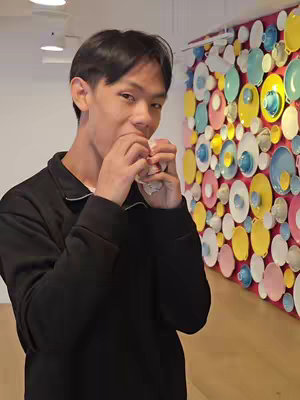 A young man with dark hair is seen in a medium shot, holding food in his hands and about to take a bite. He is wearing a black top. In the background, there's a wall adorned with colorful plates and cups.
