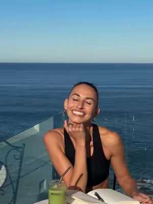 A woman is sitting at a table outdoors on a balcony, enjoying a sunny day. The background shows a clear blue sky meeting the ocean. The woman is smiling with her eyes closed, resting her chin on her hand. On the table, there's a glass of green liquid with a straw, an open notebook with a pen, and a smartphone. She is wearing a black sports bra.