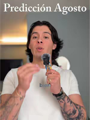 A man with dark hair and a white t-shirt is holding a microphone and gesturing with his left hand. The background is a blurred room with gray walls.