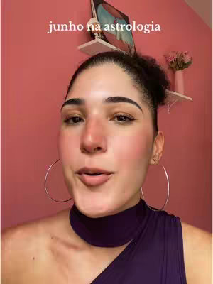 A woman with dark hair and hoop earrings is centered, facing the camera. She's wearing a purple top. The background is a pink wall, and there is text at the top of the image.