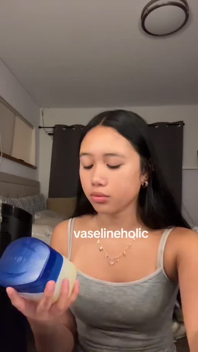 A medium shot shows a fair-skinned, young Asian woman in a tank top. She is holding a large container of Vaseline petroleum jelly, looking down at it with a neutral expression. The background is a room with a textured wall and a curtain. The lighting seems to come from the ceiling, casting slight shadows.