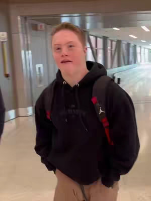 A candid shot of a young man with Down syndrome walking through a well-lit airport hallway. He wears a black hoodie, a backpack, and looks directly at viewer.