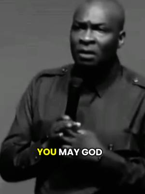 A grayscale image features a bald man speaking into a microphone. Text overlayed at the bottom of the image reads, 'YOU MAY GOD'. The image has a medium shot composition, with the man's upper body and head visible. The background is a plain, out-of-focus gray.